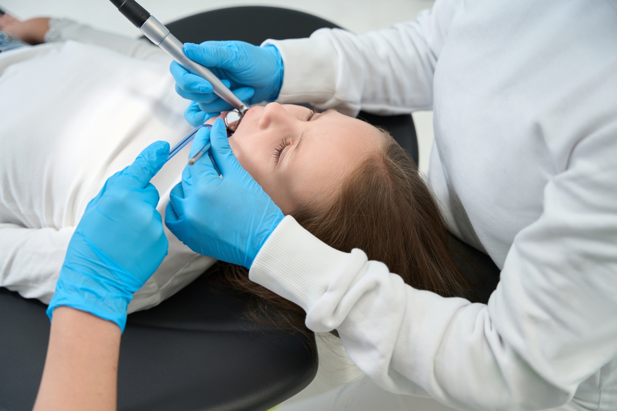 Pediatric dentist treating caries in preteen child aided by assistant